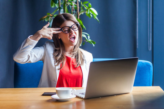 Portrait of crazy panic beautiful stylish brunette young woman in glasses sitting with pistol hand gun on her head and sceaming. indoor studio shot, cafe, office background.