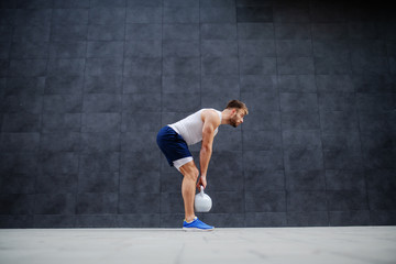 Fototapeta na wymiar Strong muscular handsome Caucasian man in shorts and t-shirt standing outdoors and lifting kettle bell. In background is gray wall.