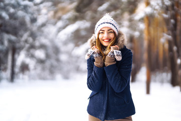 Portrait of young beautiful girl with long hair, in a blue jacket and knitted hat and mittens posing in sunny winter day. Fashion young woman in the winter forest. Christmas, winter holidays concept. 