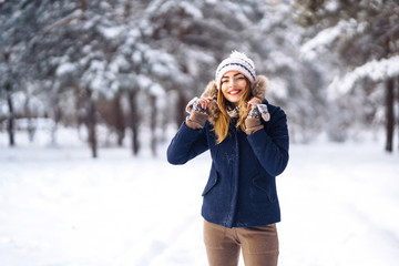 Portrait of young beautiful girl with long hair, in a blue jacket and knitted hat and mittens posing in sunny winter day. Fashion young woman in the winter forest. Christmas, winter holidays concept. 