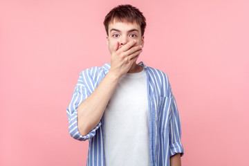 I won't tell. Portrait of amazed scared brown-haired man in casual striped shirt covering mouth with hand, keeping secret, terrified with shocking news. indoor studio shot isolated on pink background