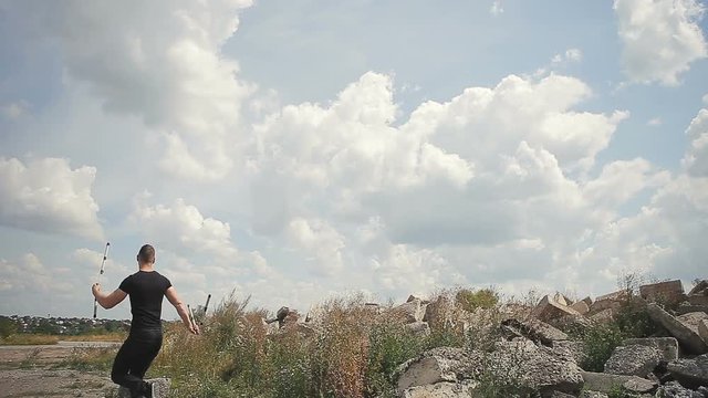 Handsome, young man with a beard in a black T-shirt demonstrates mastery with nunchaku outdoors on a stone background.