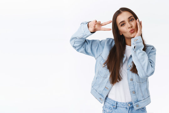 Flirty and cute, coquettish sassy woman in trendy outfit, touching cheek tilting head feminine, showing peace or victory sign near eye as sending positive vibes to audience, white background