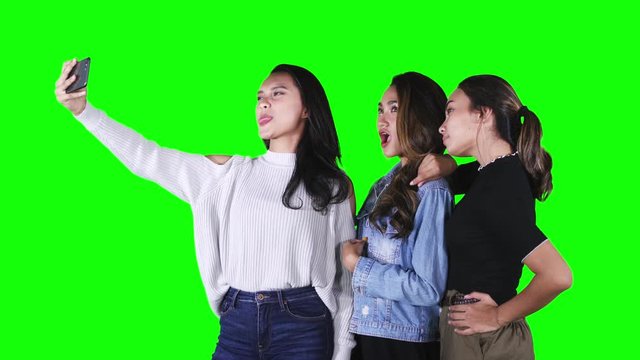 Group of three pretty teenage girls taking selfie picture together with a mobile phone in the studio. Shot in 4k resolution with green screen background