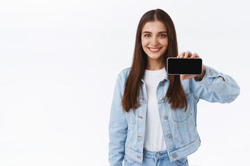 Confident cute brunette female in denim jacket, holding smartphone horizontally, promoting telephone online game or application, showing mobile screen, smiling pleased, white background
