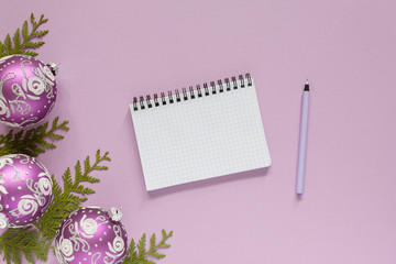 Holiday background , lilac christmas balls and thuja twigs on a pink background , open spiral notepad and pen, flat lay, top view