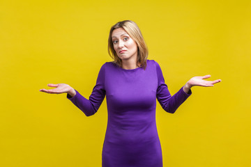 I don't know, whatever. Portrait of uncertain confused woman in tight purple dress standing with raised hands, shrugging shoulders in bewilderment. indoor studio shot isolated on yellow background