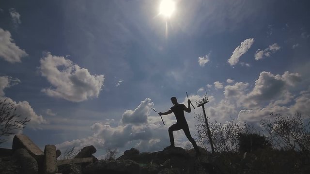 Handsome, young man with a beard in a black T-shirt demonstrates mastery with nunchaku against the sky background.