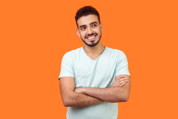 Portrait of dreamy thoughtful bearded man with toothy smile in white t-shirt standing with crossed hands, dreaming and thinking of pleasant memories. indoor studio shot isolated on orange background