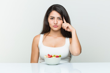 Young hispanic woman eating a fruit bowl pointing his temple with finger, thinking, focused on a task.