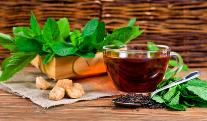 Cup of black tea with dried tea in spoon, brown sugar and fresh mint leaves in basket on a wooden background.
