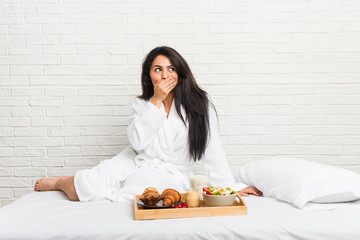 Young curvy woman taking a breakfast on the bed thoughtful looking to a copy space covering mouth with hand.