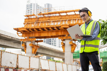 Image outside the industrial construction engineers in yellow protective ear muff discuss new project while using laptop and happy smile on the open building site near the crane.