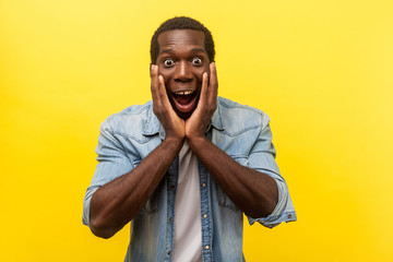 Oh my god, wow! Portrait of stunned shocked man in denim shirt holding hands on face and looking with big amazed eyes, surprised by unbelievable news. indoor studio shot isolated on yellow background