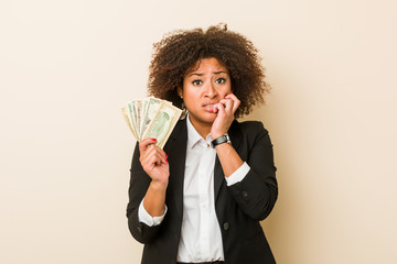 Young african american woman holding dollars biting fingernails, nervous and very anxious.