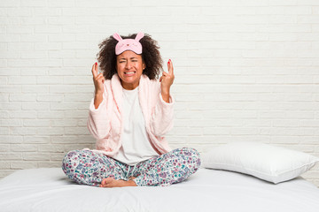 Young african american woman in the bed wearing pijama crossing fingers for having luck