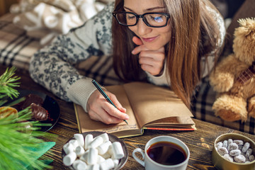 Girl in a sweater is writing a wish list in a notebook in the warm atmosphere of Christmas. Cozy New Year mood - obrazy, fototapety, plakaty