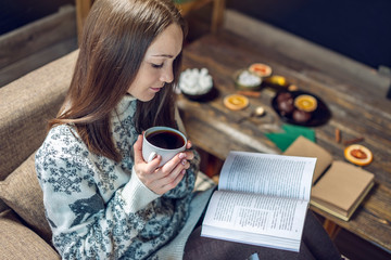 Woman in a sweater reading a book with a mug of coffee in the evening in a warm Christmas...