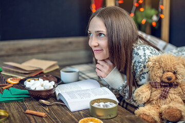 Young girl in a sweater reading a book with cup of coffee in the evening in a warm Christmas...