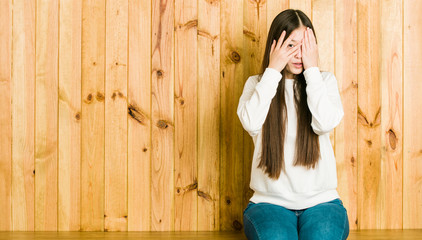Obraz premium Young chinese woman sitting on a wooden place blink through fingers frightened and nervous.