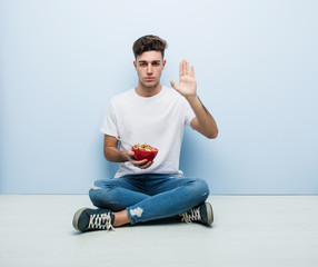Young man eating cereals sitting on the floor standing with outstretched hand showing stop sign,...