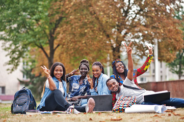 Group of five african college students spending time together on campus at university yard. Black afro friends sitting on grass and studying with laptops.