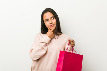Young hispanic woman holding a shopping bag looking sideways with doubtful and skeptical expression.