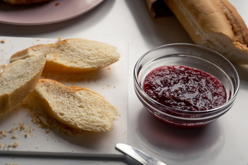 A glass bowl with sweet raspberry jam, slices of baguette on a white table in the kitchen in the morning. 