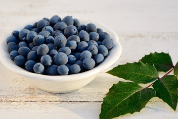 Mahonia berries in a plate and leaves on a white wooden background close up