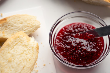 A knife in a glass bowl with sweet raspberry jam, and slices of baguette for breakfast on a white table in the kitchen in the morning. 