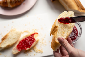 A woman begins to spread with the knife a vitamin C raspberry jam on a fresh sliced ​​baguette for breakfast over a white table in the kitchen. Close-up.