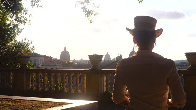 Young woman tourist in fashion white dress with hat walking at panoramic view of Rome cityscape from campidoglio terrace at sunset. Landmarks, domes of Rome, Italy.