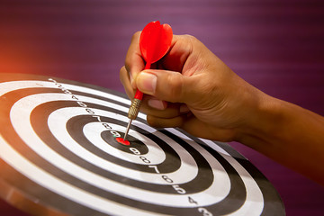 Hand holding red dart arrow  hitting in the target center of dartboard on bullseye.