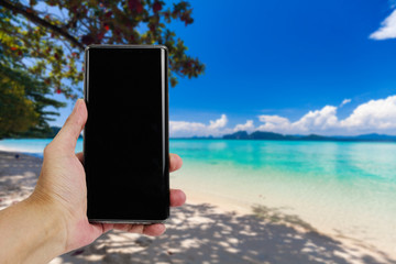 Hand hold mobile phone on the beach with beautiful sea and blue sky background. Beach holiday concept.