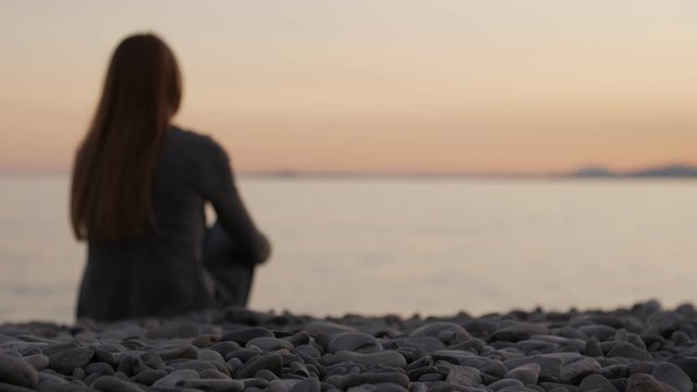 Silhouette of a Girl Sitting on the Beach at Sunset. Romantic scene lonely woman on the beach in the evening. Focused on pebbles in the foreground.