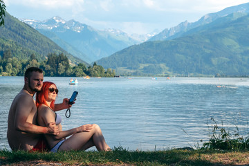 A young couple sits on the lake communicate with each other, uses a smartphone, enjoy the views, vacation, relax. couple in love Zell am see in Austria, copy space, selective focus, summer day