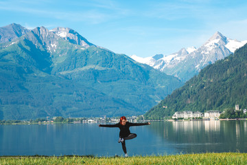 An attractive young woman doing a yoga pose for balance and stretching near the lake Zell am See high in the Alpine mountains in Austria. dressed in a black tracksuit for training
