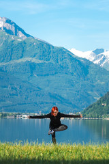 Fototapeta na wymiar An attractive young woman doing a yoga pose for balance and stretching near the lake Zell am See high in the Alpine mountains in Austria. dressed in a black tracksuit for training, vertical photo