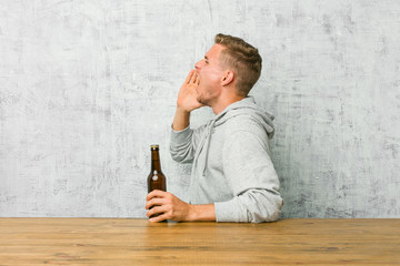 Young man drinking a beer on a table shouting and holding palm near opened mouth.