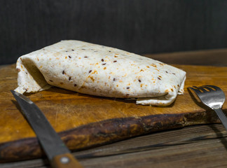 Classic tortilla wrap on cutting board lying on wooden table with fork and knife with black background.