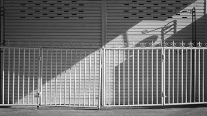 Sunlight and shadow on surface of white metal fence with vintage black hanging pole in front of the old shutter door background in morning time, black and white style