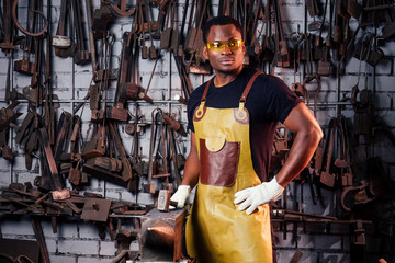 handsome african americam man forging steel next to furnace in dark workshop. small business...