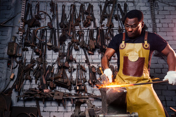 Fototapeta na wymiar handsome african americam man forging steel next to furnace in dark workshop. small business comcept