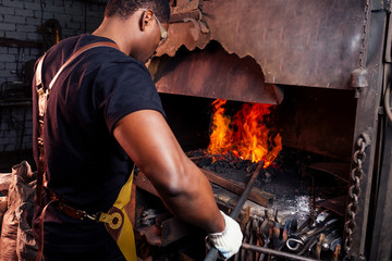 handsome african americam man forging steel next to furnace in dark workshop. small business comcept