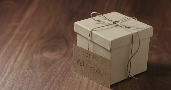 Slow motion man hand put happy new year card next to brown paper gift box on walnut table