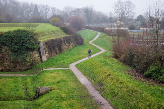 Misty winter day. A winding path passes near the earthen fortifications of an ancient fortress in the city of Bergues, France. The silhouette of a passing cyclist is visible. Background.