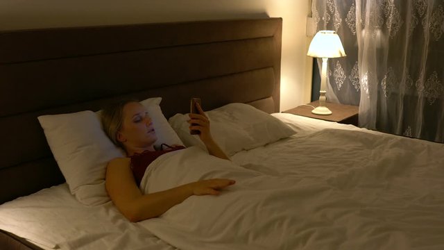 young woman lies in bed in the company of a cat and uses a smartphone