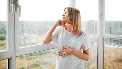 Portrait of a pregnant happy woman standing at panoramic window on a fresh rainy autumn day, smiling at her thoughts.