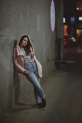 Naklejka premium A beautiful girl in a pink sweater with a hood stands against the backdrop of the night city with a smartphone in her hand. Soft focus, colored highlights in the background.
