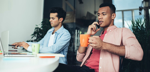 African American male employee calling to operator for consultancy about 4g wireless connection during coffee time at office desktop, two skilled programmers working hard in coworking space